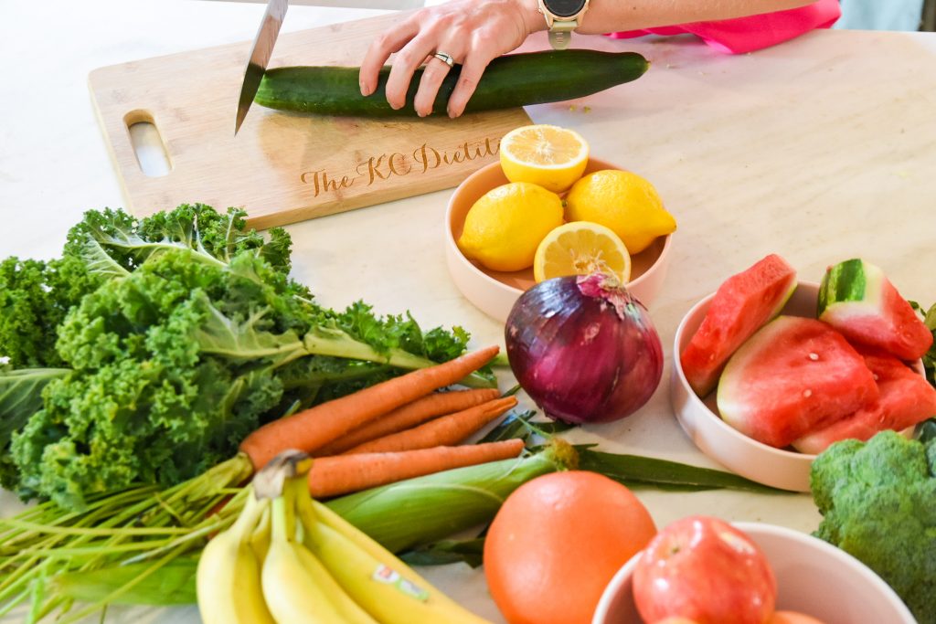 fruits and vegetables on a table with a hand cutting a cucumber on an engraved wooden cutting board in the background