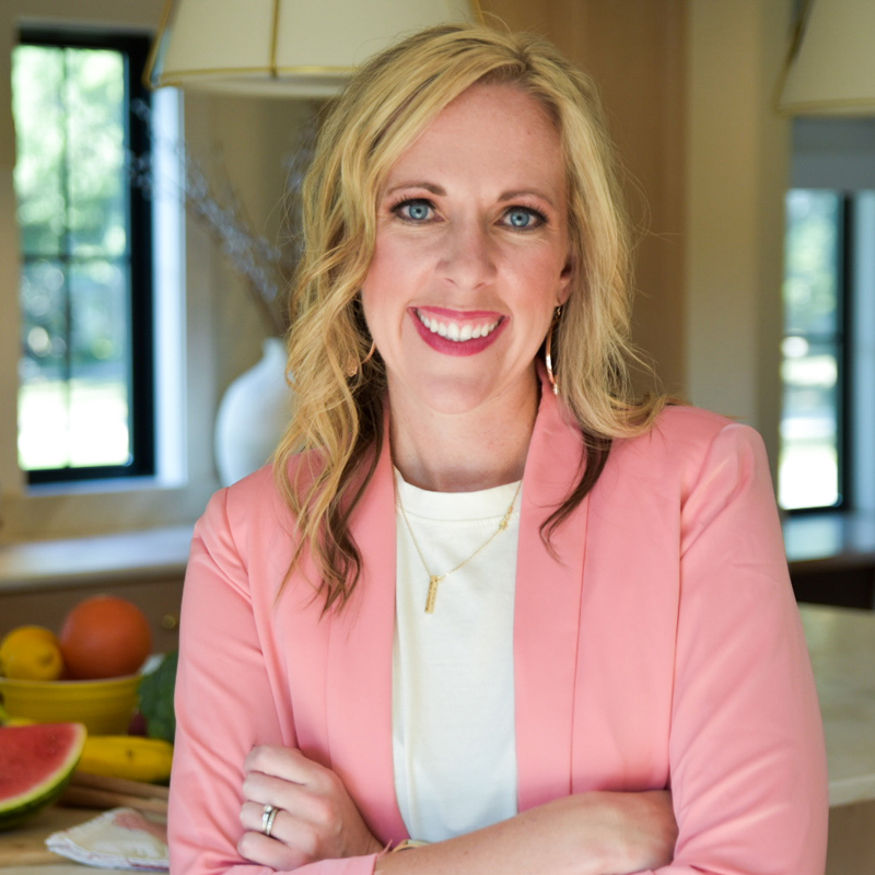 smiling woman with blonde hair, blue eyes, and crossed arms wearing gold hoop earrings and necklace, and a white shirt under a pink blazer