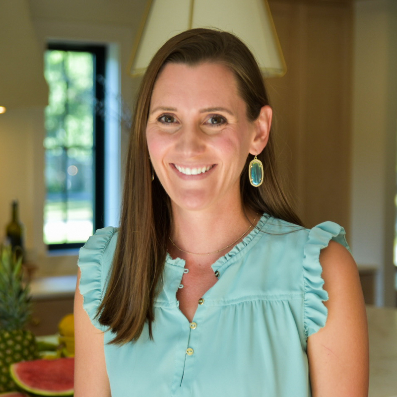 smiling woman with long, brown, side-parted hair wearing blue and gold dangly earrings, a necklace, and a blue tank blouse with gold buttons