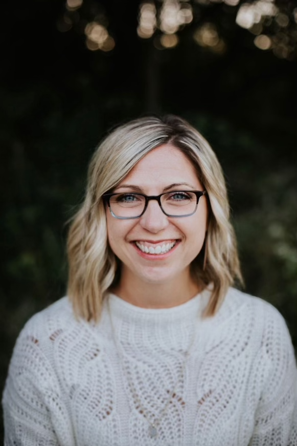 white woman smiling wearing white cable knit sweater, glasses, and dirty blonde shoulder-length hair parted to one side